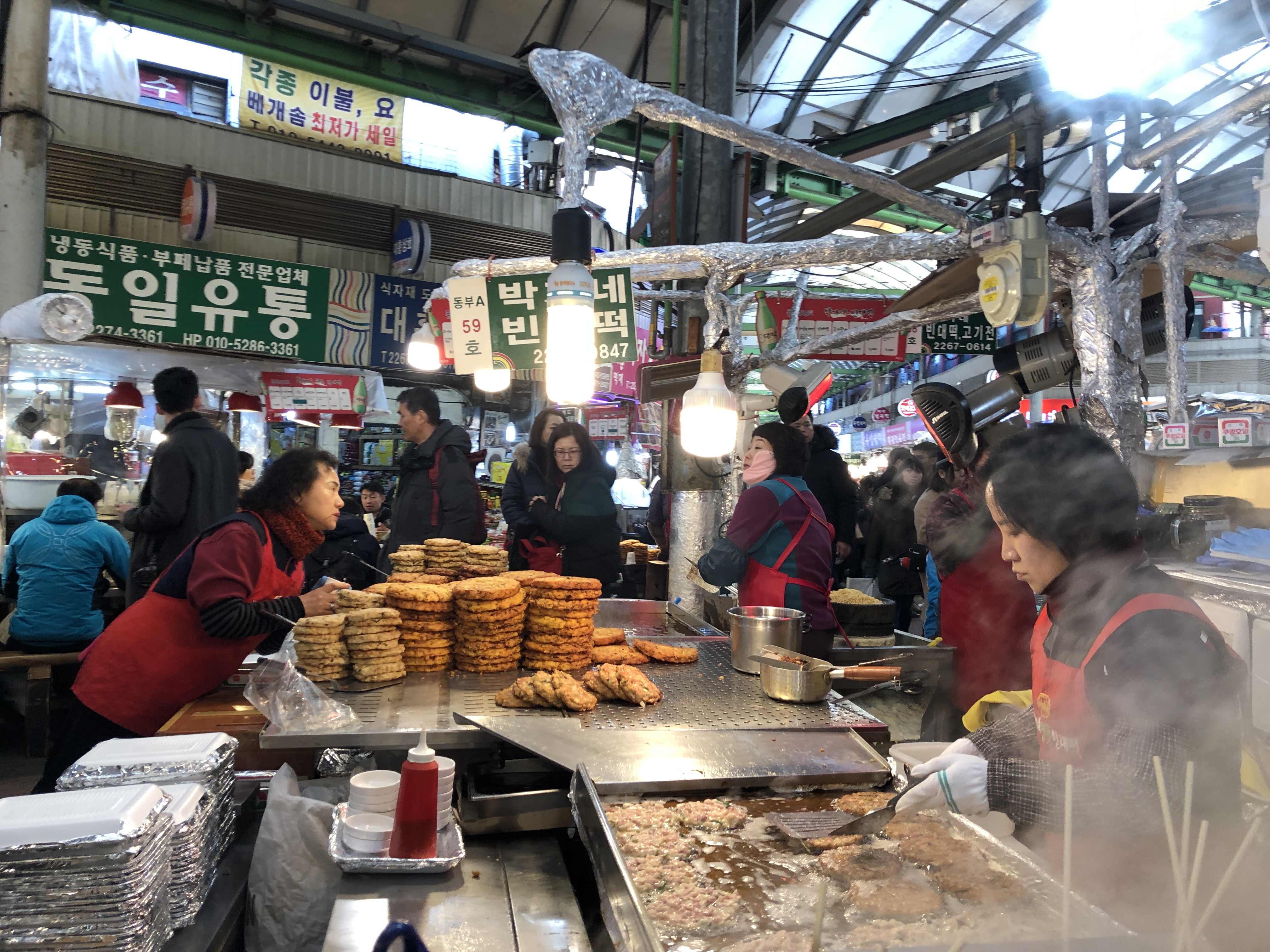 Gwangjang Market in Seoul, Korea. Photo of open air food market with several food stalls and vendors selling to the crowds. Large table has multiple stacks of mung bean pancakes.