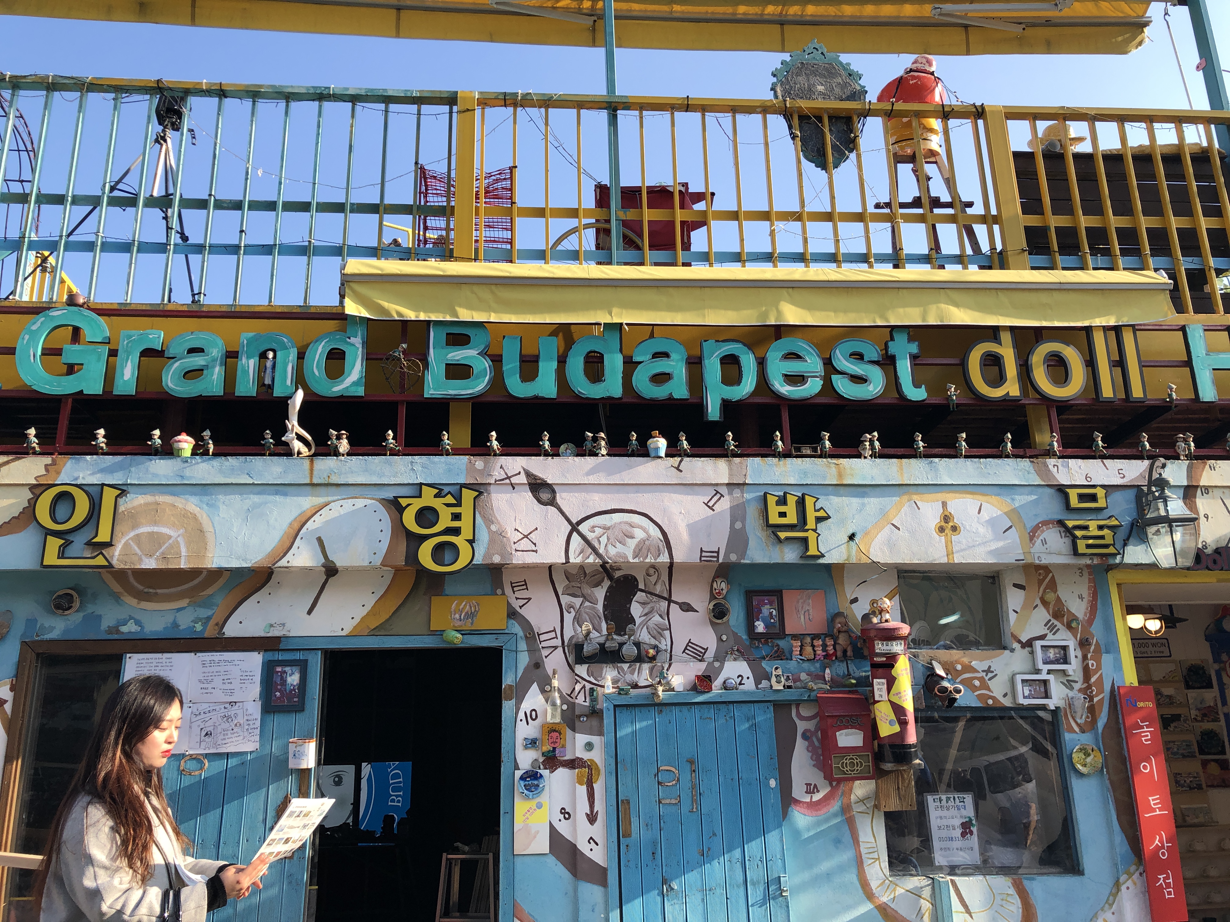 Front of Grand Budapest Doll Hotel in Gamcheon Cultural Village in Busan, Korea. Front is covered in art. Woman is reading a paper. 